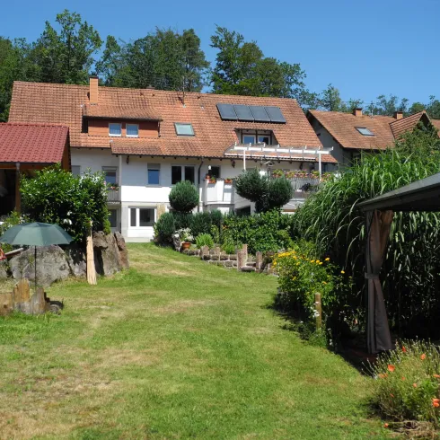 Ein gepflegter Garten mit grünen Wiesen und mehreren Bäumen. Im Hintergrund sind gemütliche Wohnhäuser unter klarem blauen Himmel zu sehen.