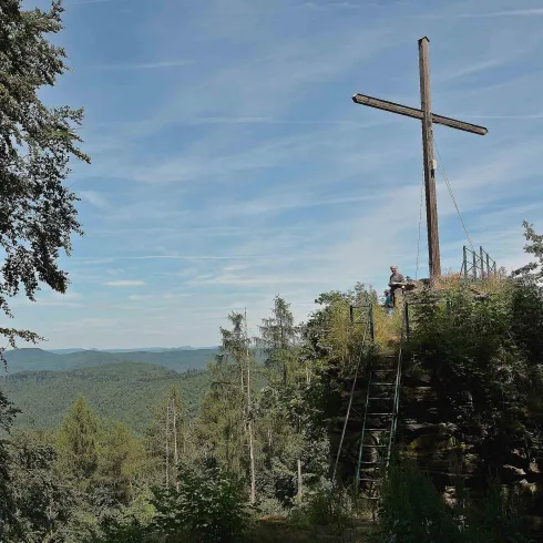 Ein großes Holzkreuz steht auf einem Hügel, umgeben von Bäumen. Im Hintergrund erstreckt sich eine Aussicht auf sanfte Hügel und einen klaren Himmel.