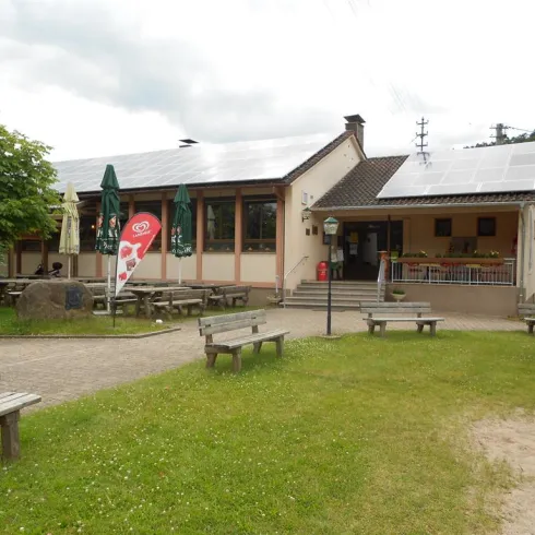 Ein gemütliches Gebäude mit mehreren Fenstern und Sonnenschirmen. Im Vordergrund sind Bänke auf einer Wiese und ein Eis-Stand.