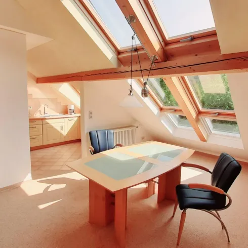Ein helles, modernes Zimmer mit Dachfenstern und einem gemütlichen Tisch. Die Einrichtung ist schlicht und einladend, ideal für Arbeiten oder Entspannen.