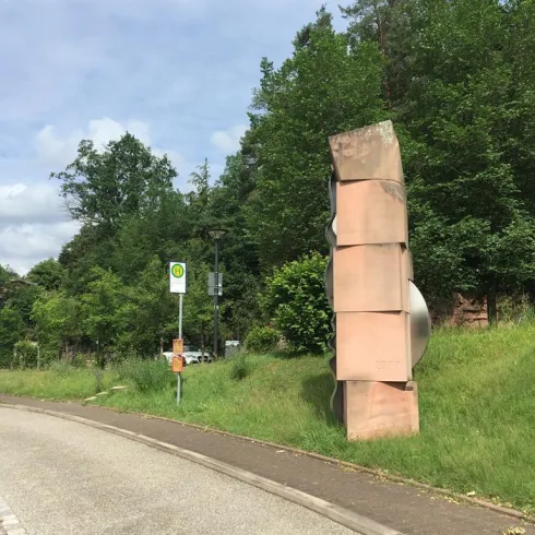 Eine künstlerische Steinskulptur steht an einer Straße, umgeben von Bäumen. Im Hintergrund sind Verkehrsschilder und eine bushaltestelle sichtbar.