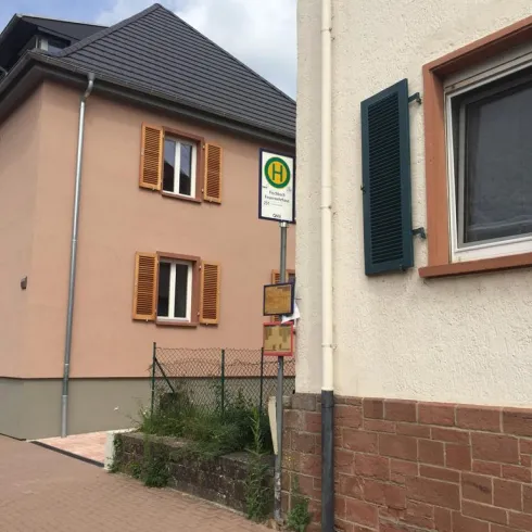 Eine ruhige Straßenansicht mit zwei Gebäuden. An einer Wand befindet sich ein Bushaltestellenschild.