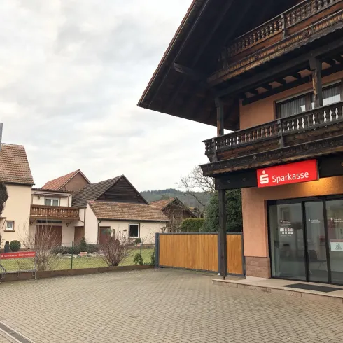 Ein modernes Bankgebäude mit dem Logo der Sparkasse. Im Hintergrund sind Wohnhäuser und ein ruhiger Platz zu sehen.