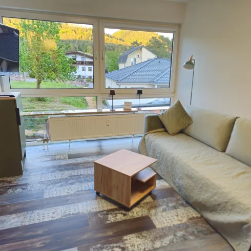 Ein gemütliches Wohnzimmer mit einem bequemen Sofa und einem kleinen Holzkaffee-Tisch. Große Fenster bieten einen Blick auf die umliegende Natur und die Nachbarhäuser.