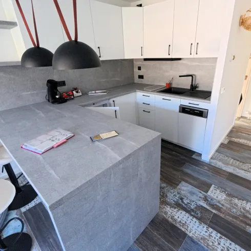 Eine moderne Küchenzeile mit grauem Arbeitsbereich und weißen Schränken. Im Hintergrund sind stylische Barhocker zu sehen.