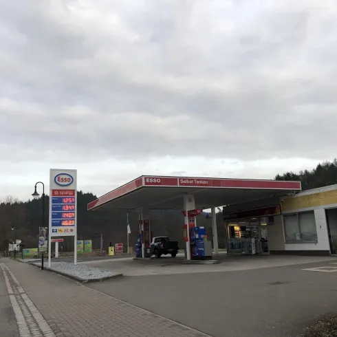 Eine Esso-Tankstelle mit einer Zapfsäule und einem benachbarten Geschäft. Der Himmel ist bewölkt und die Umgebung ist ruhig.