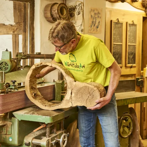 Ein Mann in einem gelben T-Shirt arbeitet in einer Holzwerkstatt. Er hält ein kunstvoll geformtes Holzstück und steht vor einer Holzmaschine.