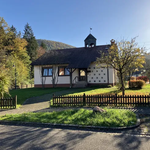 Ein charmantes Haus mit einem steilen Dach und einer kleinen Glocke. Der Garten ist grün und gepflegt, umgeben von Bäumen und einem Zaun.