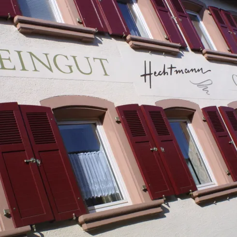 Ein Weingut mit dem Namen "Hechtmann" ist an einer Wand zu sehen. Die Fenster haben rote Fensterläden und weiße Vorhänge.