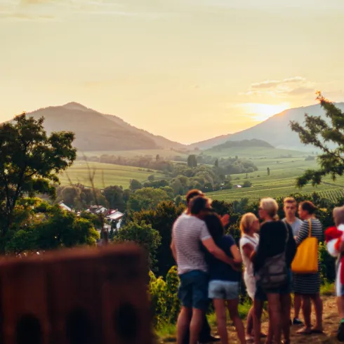 Eine Gruppe von Menschen steht in einer malerischen Landschaft mit sanften Hügeln im Hintergrund. Der Himmel ist in warmen Farben während des Sonnenuntergangs gefärbt.
