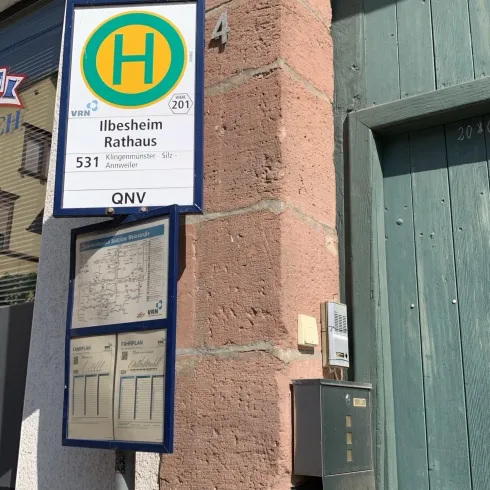 Eine Bushaltestelle mit dem Schild für "Ilbesheim Rathaus" ist zu sehen. Daneben befindet sich ein Briefkasten und eine Tür.