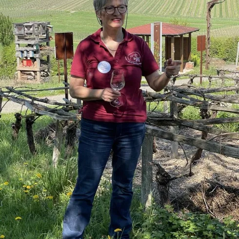 Eine Frau steht in einem Weinberg und hält ein Weinglas in der Hand. Im Hintergrund sind grüne Weinreben und sanfte Hügel zu sehen.