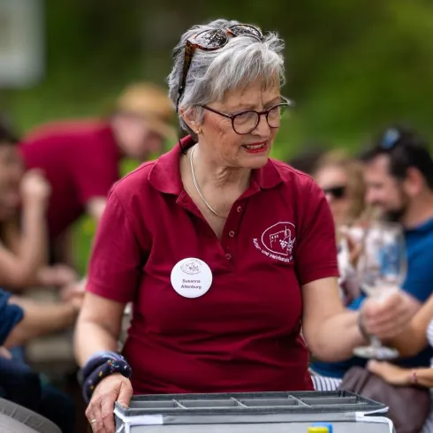 Eine ältere Dame in einem roten Polohemd serviert Getränke bei einer Veranstaltung im Freien. Im Hintergrund sind viele Menschen, die miteinander reden und die Atmosphäre genießen.