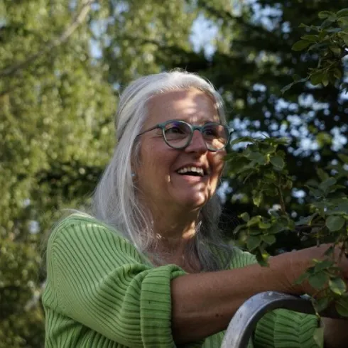 Eine lächelnde Frau mit langen grauen Haaren arbeitet in einem Garten. Sie trägt ein grünes Oberteil und sieht glücklich aus.