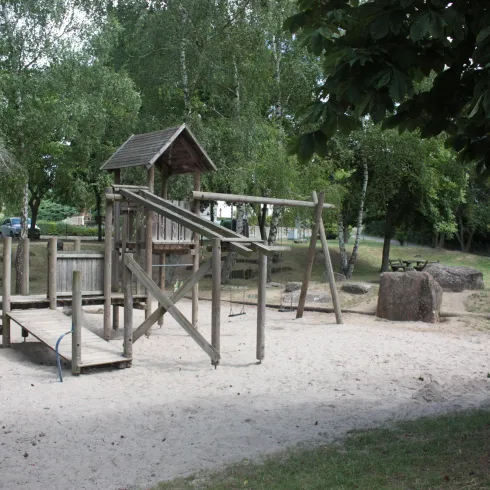 Ein Spielplatz aus Holz mit einer Rutsche und einer Schaukel. Die Umgebung ist grün mit vielen Bäumen.