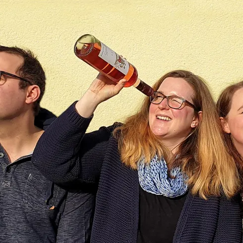 Drei Personen halten Weinflaschen ans Ohr, als ob sie lauschen. Sie stehen vor einer gelben Wand und lächeln.