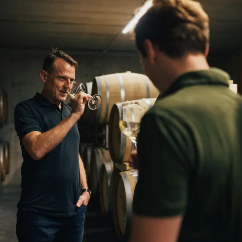 Zwei Männer stehen in einem Weinkeller mit Holzfässern. Einer der Männer riecht an einem Glas Wein, während der andere aufmerksam zuhört.