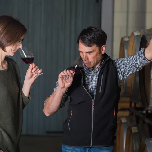 Ein Mann und eine Frau verkosten Rotwein in einem Weinkeller. Im Hintergrund sind Holzfässer zu sehen.