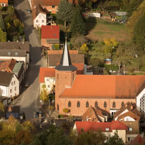Eine Ansicht eines kleinen Dorfes mit Häusern und einer Kirche. Die Kirche hat einen markanten Turm und ist von bunten Bäumen umgeben.