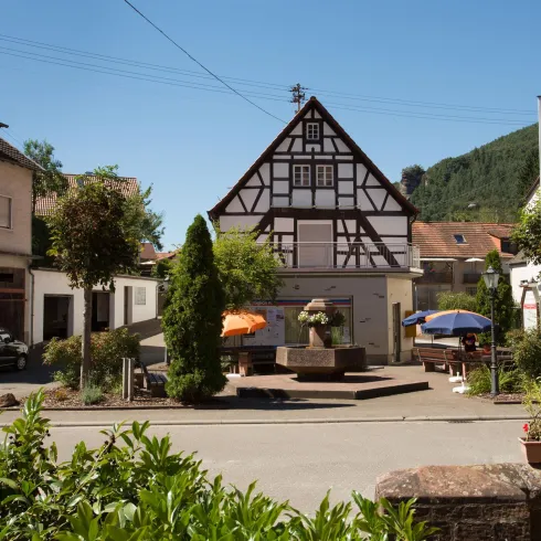 Dorfbrunnen in Lug. Dahinter im Fachwerkgebäude befindet sich ein kleines Geschäft mit Backwaren etc.