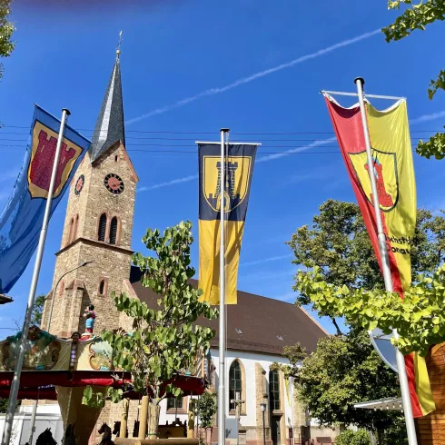 Bunte Fahnen wehen an Fahnenmasten. Im Hintergrund die Kirchturmspitze von Ruppertsberg. 