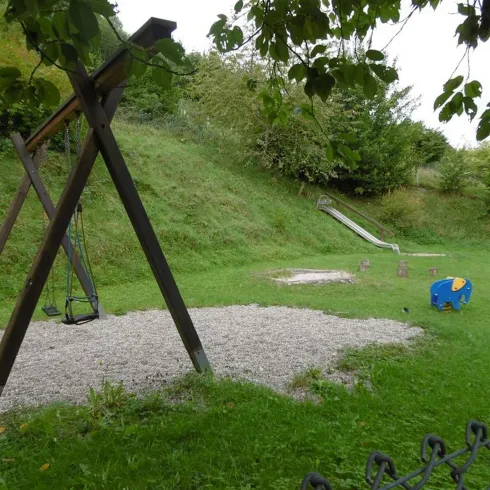 Ein Spielplatz mit einer Schaukel und einer Rutsche ist zu sehen. Im Vordergrund gibt es einen bunten Spielzeug-Elefanten und einen kiesbedeckten Bereich.