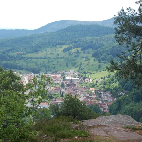 Eine malerische Aussicht auf ein kleines Dorf im Tal, umgeben von grünen Hügeln. Der Himmel ist bewölkt und die Landschaft wirkt friedlich.