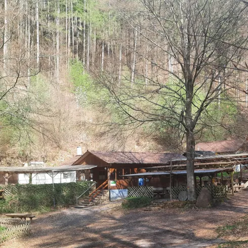 Ein einfaches Holzgebäude im Wald, umgeben von Bäumen und natürlicher Vegetation. Der Weg vor dem Gebäude ist gut erkennbar und zeigt eine ruhige, ländliche Atmosphäre.