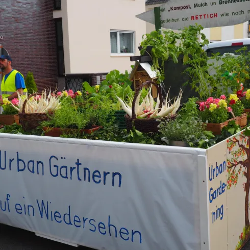 Ein Wagen mit vielen verschiedenen Pflanzen und Kräutern steht auf der Straße. Darauf steht der Schriftzug "Urban Gärtnern" und es gibt eine dekorative Baumgrafik.