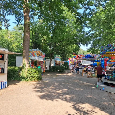 Eine belebte Festplatz mit bunten Fahrgeschäften und Ständen. Dicke Bäume spenden Schatten und schaffen eine fröhliche Atmosphäre.