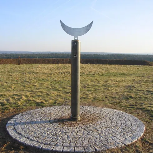 Ein metallisches Kunstwerk in Form einer Halbmondskulptur auf einem Steinfundament. Die umgebende Landschaft ist weitläufig und offen.