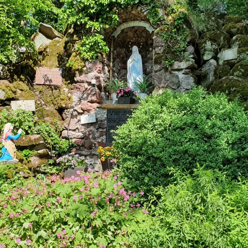Eine kleine Mariensäule ist in einer grünen Landschaft aus Büschen und Blumen eingebettet. Die Umgebung ist von üppigem Grün und Steinen geprägt.