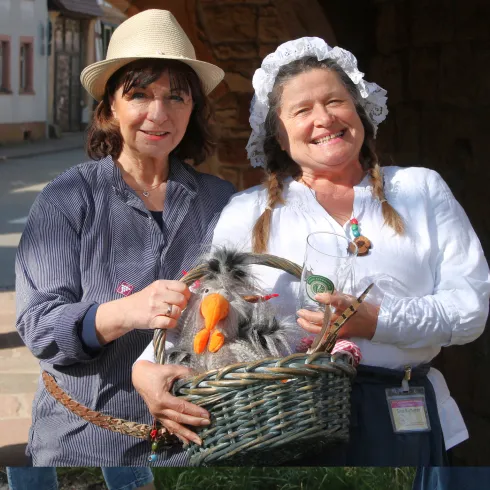 Zwei Frauen stehen in einer malerischen Straße. Sie halten einen Korb mit handgefertigten Figuren und lachen freundlich.