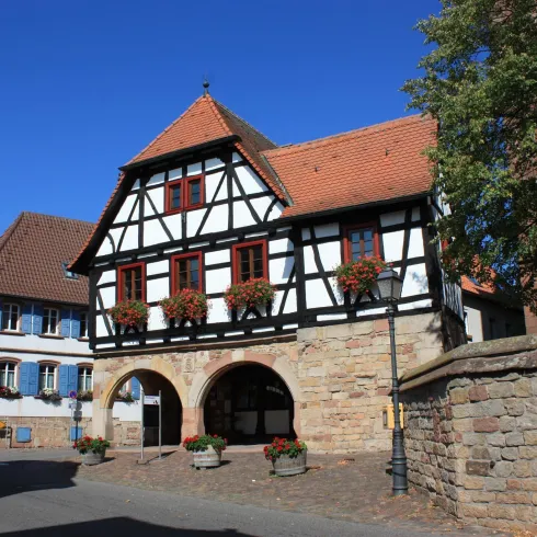 Ein historisches Fachwerkhaus mit blühenden Blumenkästen und einem roten Dach. Im Hintergrund sind weitere traditionelle Gebäude zu sehen.