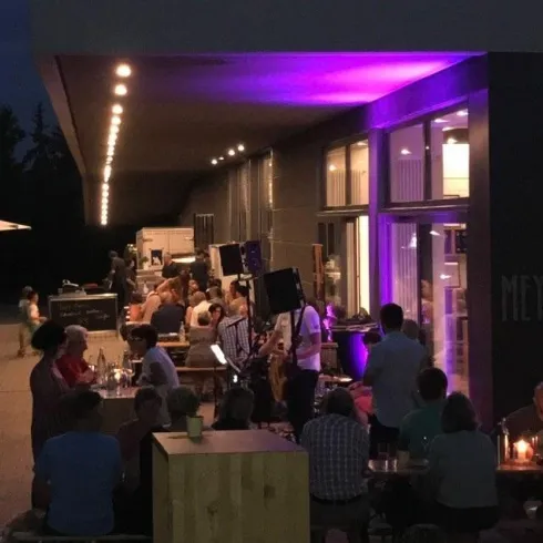 Eine belebte Bar im Freien bei Nacht, umgeben von Gästen, die entspannt sitzen. Sanftes Licht von der Bar sorgt für eine gemütliche Atmosphäre.