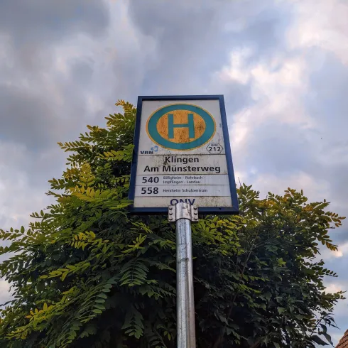 Eine Haltestelle mit einem Bus-Schild. Im Hintergrund sind Wolken und Bäume zu sehen.