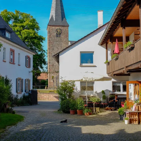 Eine malerische Gasse mit historischen Gebäuden und einer Kirche im Hintergrund. Überall sind Pflanzen und gemütliche Sitzecken zu sehen.