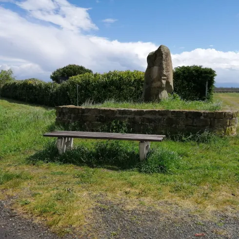 Eine Holzbank steht im Vordegrund, zusammen mit einem Weg der an ihr vorbei. Im Hintergrund ist eine kleine Mauer und ein großer Stein, genauso wie eine lange Hecke . Die Bank steht am Burgunderwanderweg.