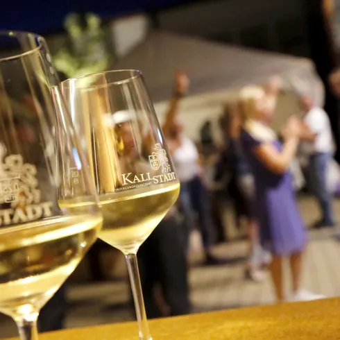 <p>Zwei Weingläser mit Aufschrift Winzergenossenschaft Kallstadt und  Gästen im Hintergrund</p>