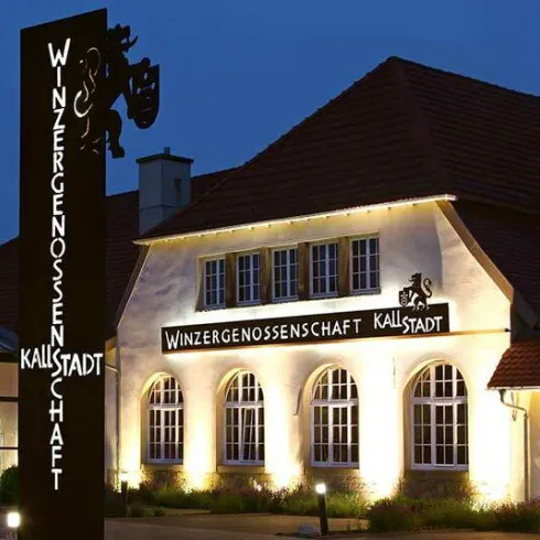 Ein schön Beleuchtetes Gebäude, die Winzergenossenschaft Kallstadt von vorne. Vor dem Gebäude steht ein großes Schild der Winzergenossenschaft.