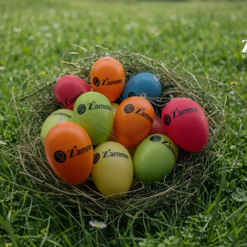 Bunte Ostereier liegen in einem Nest aus Gras. Die Eier sind mit dem Logo der Winzergenossenschaft Zamme versehen.