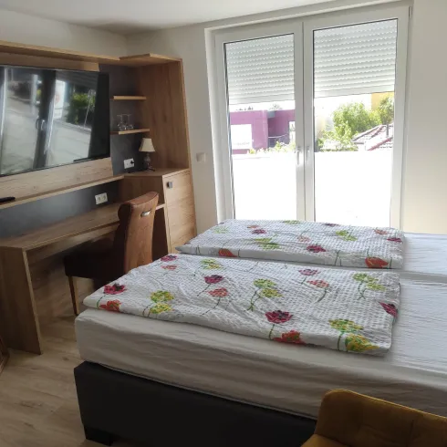 Ein modernes Schlafzimmer mit Doppelbett und einer hellen Einrichtung. Große Fenster sorgen für viel Tageslicht und bieten einen Blick nach draußen.