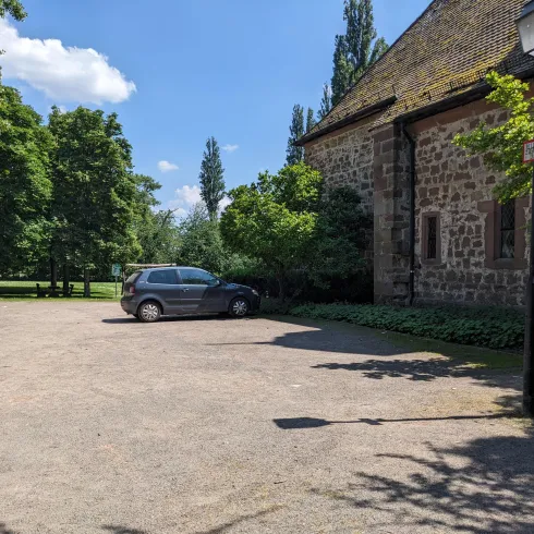 Ein ruhiger Parkplatz neben einem historischen Gebäude. Grünbäume und ein blauer Himmel schaffen eine angenehme Atmosphäre.