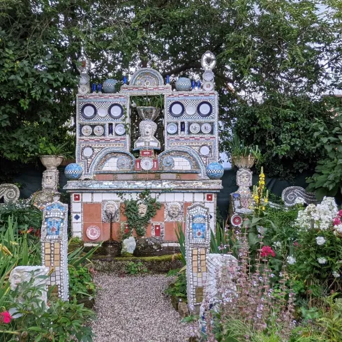 Ein farbenfrohes Grabmal, umgeben von Blumen und grüner Vegetation. Das Monument zeigt kunstvolle Details und ist von einem gepflegten Garten umgeben.