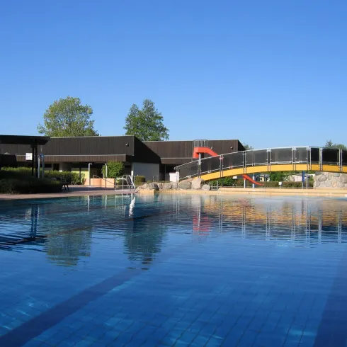 Ein ruhiger Pool mit klarem Wasser und einem blauen Himmel. Im Hintergrund befindet sich ein modernes Gebäude und eine kleine Brücke.