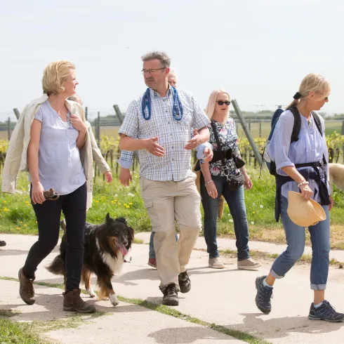 Eine Gruppe von Menschen geht auf einem Weg in der Natur. Ein Hund begleitet sie und die Stimmung wirkt entspannt und freundlich.