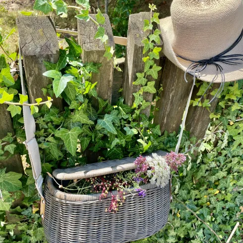 Ein Korb mit Trockenblumen hängt an einem Holzzaun, daneben liegt ein Hut. Die Umgebung ist von grünem Efeu umrahmt.