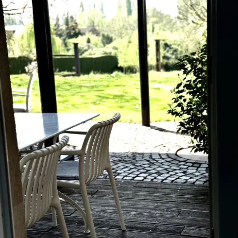 Ein schöner Blick von einem Innenraum auf eine Terrasse mit einem Tisch und Stühlen. Im Hintergrund sieht man eine grüne Wiese und Bäume.