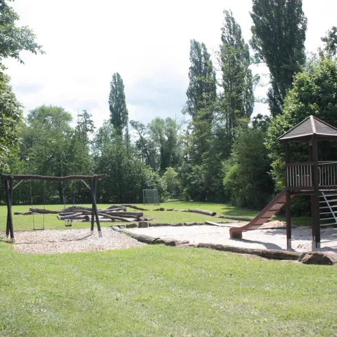 Ein Spielplatz mit einer Rutsche und einer Schaukel befindet sich in einem großen, grünen Park. Es gibt viele Bäume und einen Sandbereich zum Spielen.