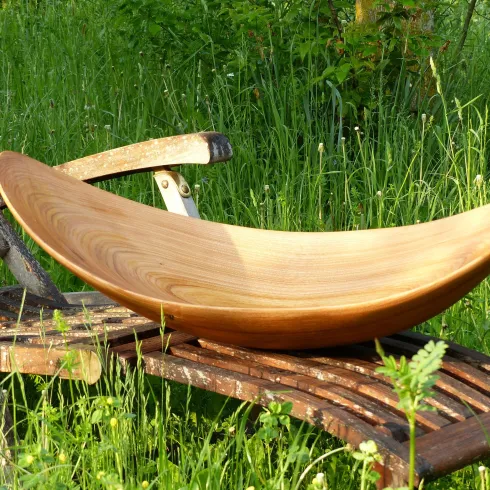Eine elegante Holzschale liegt auf einer Holzbank im Gras. Die Schale hat eine geschwungene Form und reflektiert das natürliche Licht.